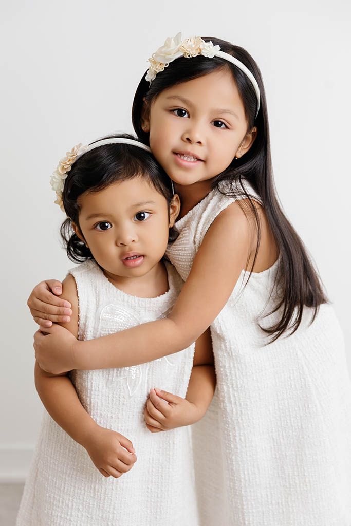 Two young sisters in matching white dresses and floral headbands hugging in a studio portrait.