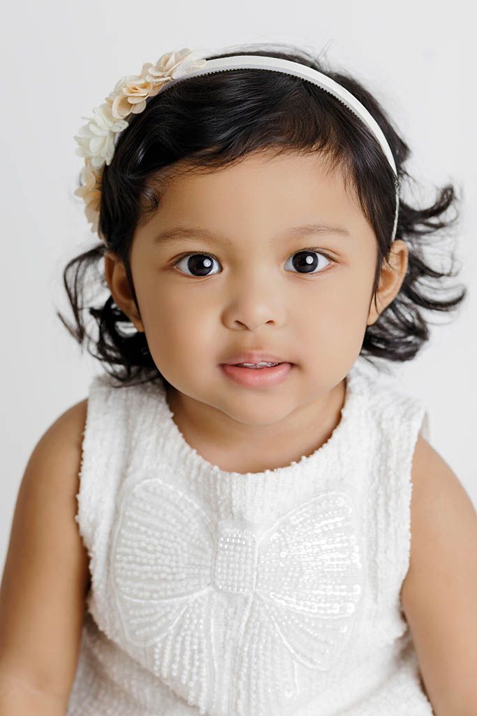 An adorable toddler girl with dark hair wearing a white bow dress and floral headband poses for a portrait.