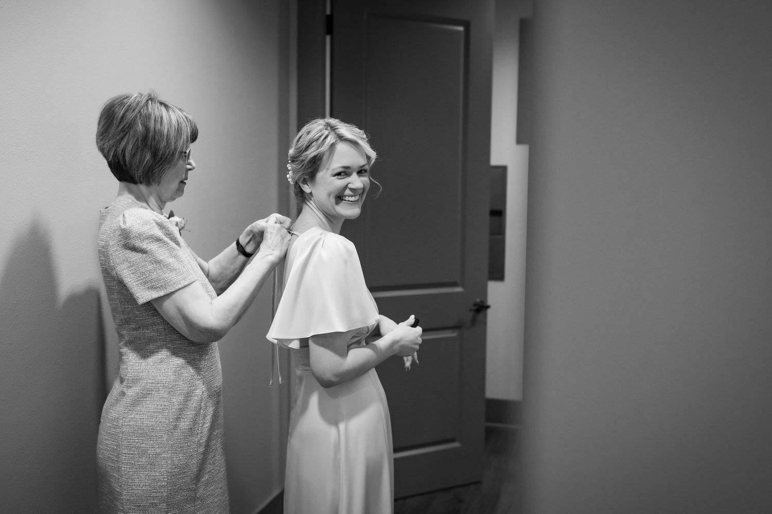 Black and white photo of a bride smiling as a woman helps adjust her wedding dress in a hallway.