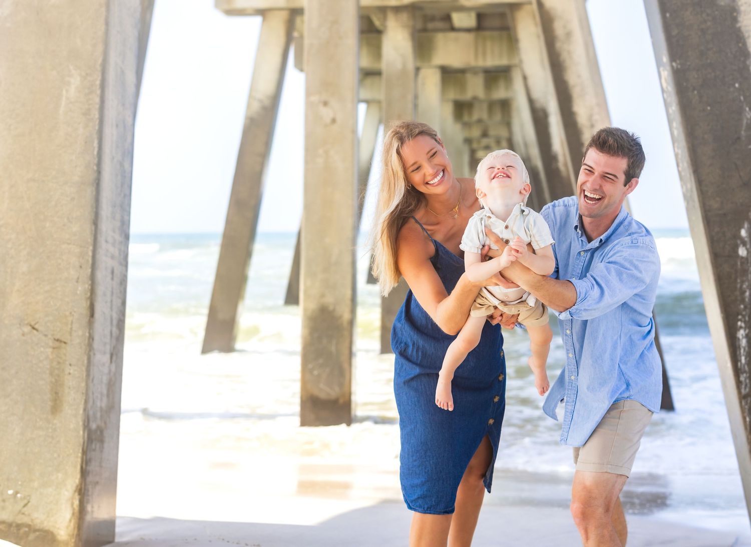 Happy young family with baby laughing together on a sunny beach near a wooden pier.