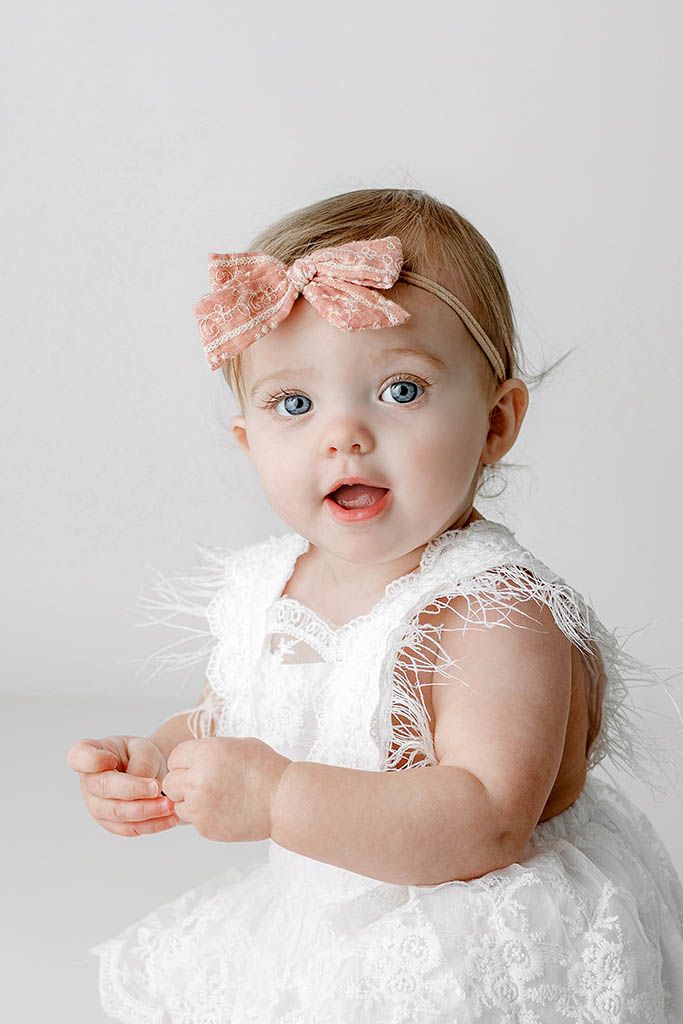 Adorable baby girl with blue eyes wearing a white lace dress and pink bow headband, sitting against a white background.