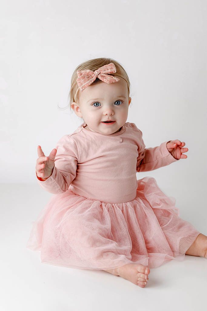 Baby girl in pink tutu dress and bow headband sits on white background, looking up with blue eyes.