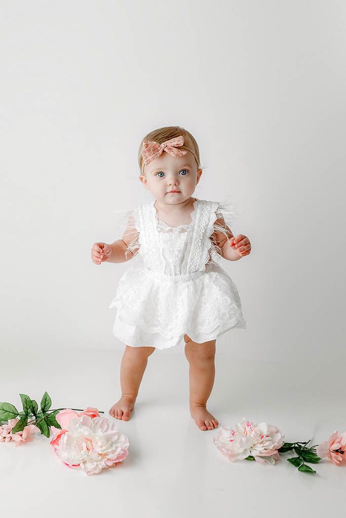 Adorable baby girl in white lace romper stands surrounded by pink peony flowers in studio portrait.