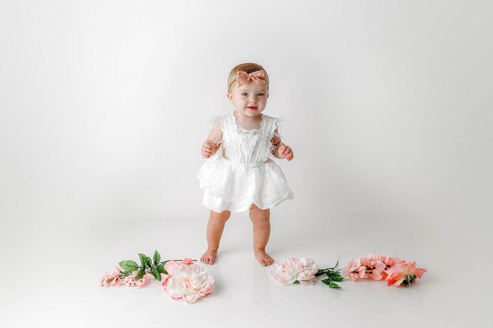 Smiling baby girl in white ruffled romper standing with pink flowers on white backdrop.