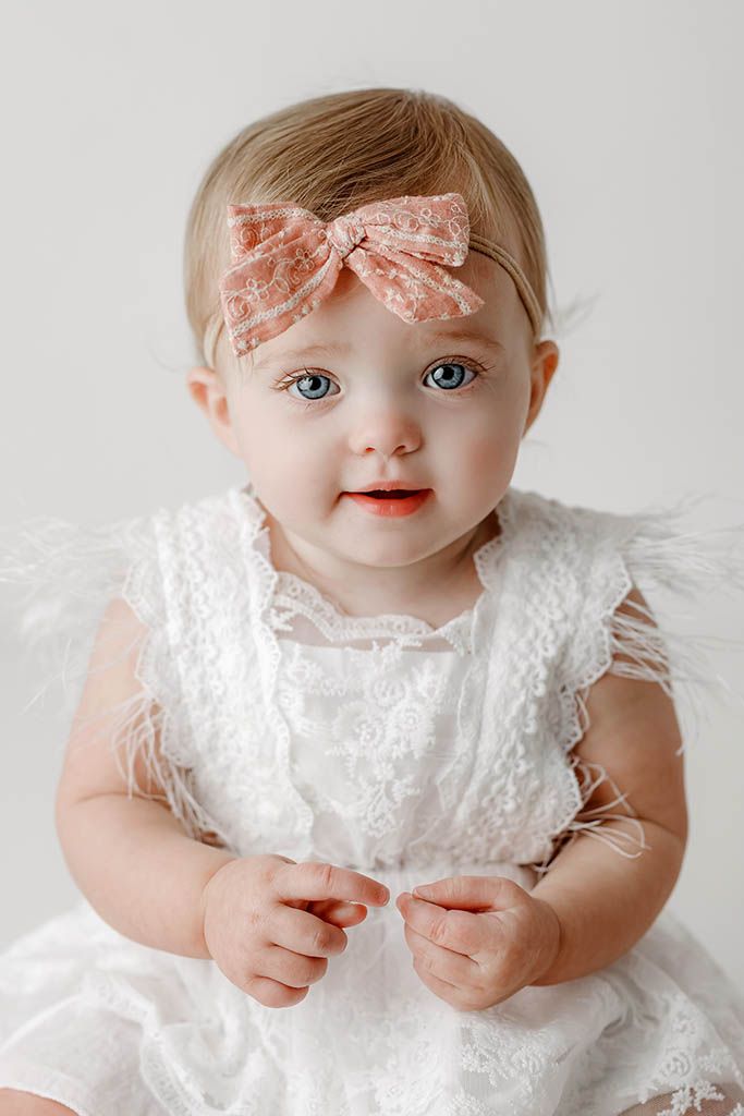 Adorable baby girl with blue eyes wearing white lace dress and pink bow headband.
