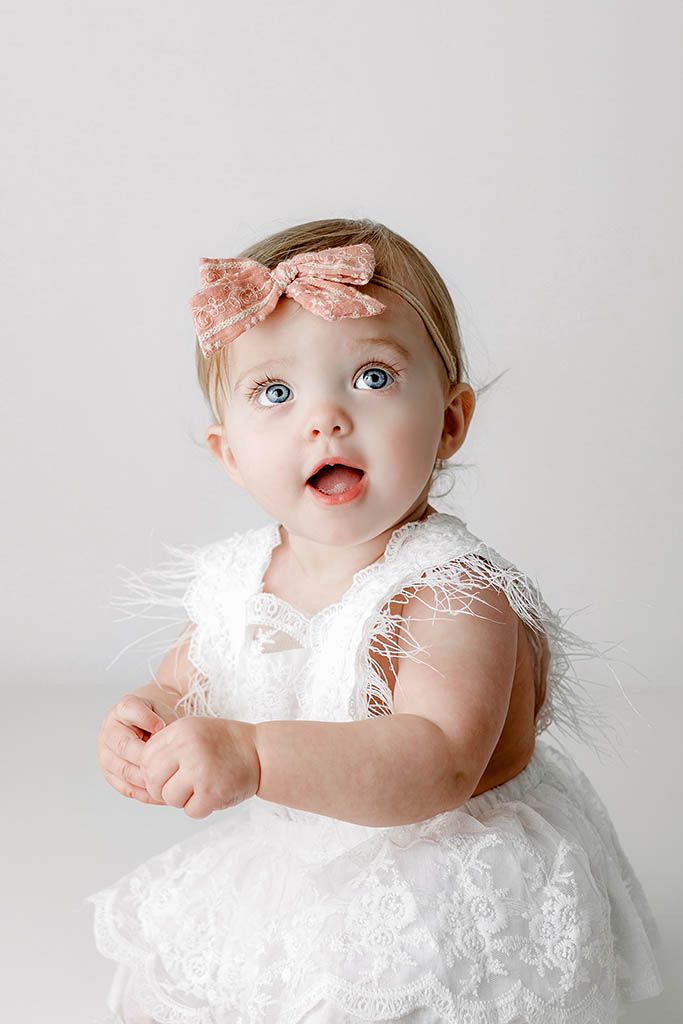 Adorable baby girl with blue eyes wearing a white lace dress and pink bow headband, sitting against a white background.