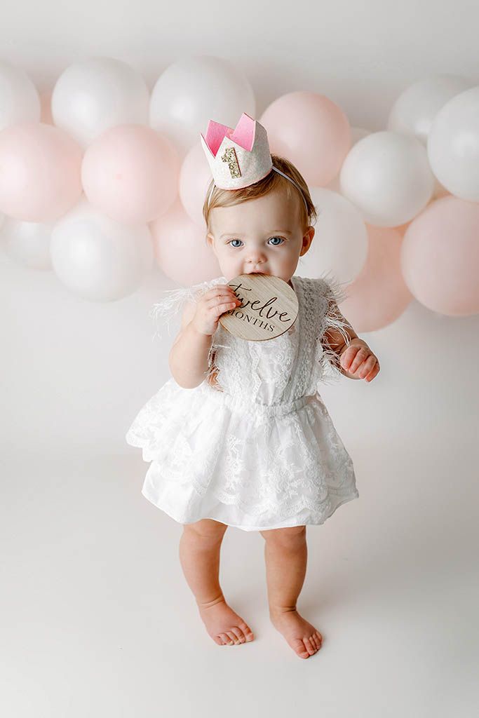 Baby girl in white dress and crown holds 12-month milestone disc, surrounded by pink and white balloons.