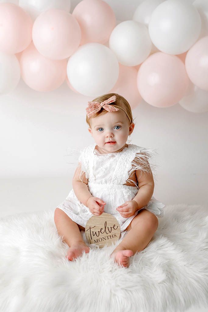 Baby girl in white feather romper holds a twelve months milestone sign, sitting on fur rug with pink and white balloons.