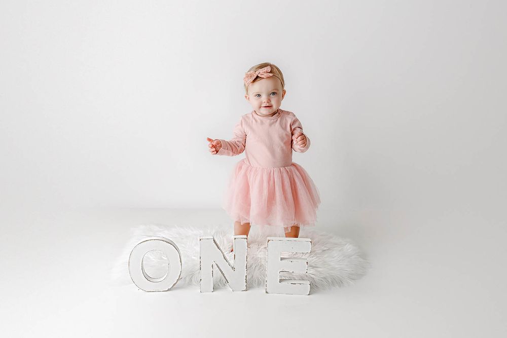 Baby girl in pink tutu dress stands on white ONE letter blocks for first birthday photo session.