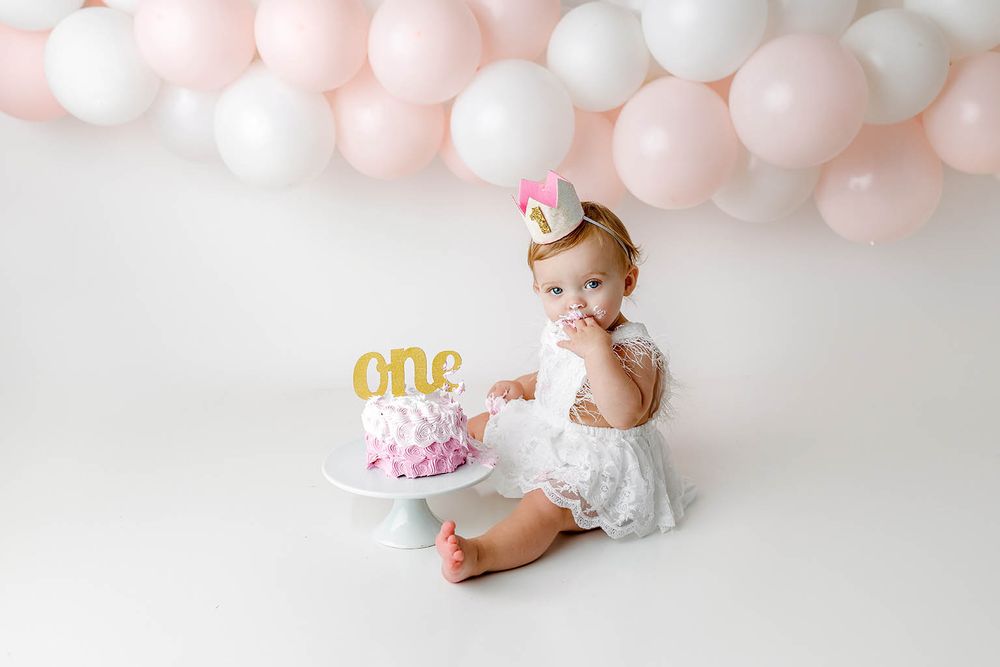 Baby girl in white dress celebrating first birthday with pink cake and balloon backdrop.