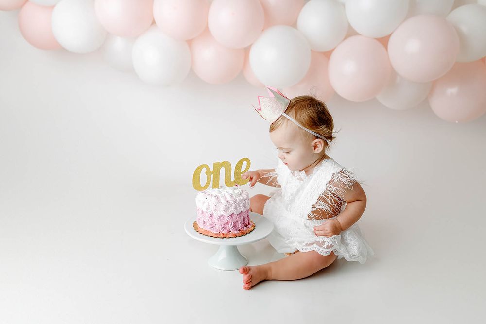 One-year-old baby girl in white dress smashes pink birthday cake with gold one topper, surrounded by pink balloons.