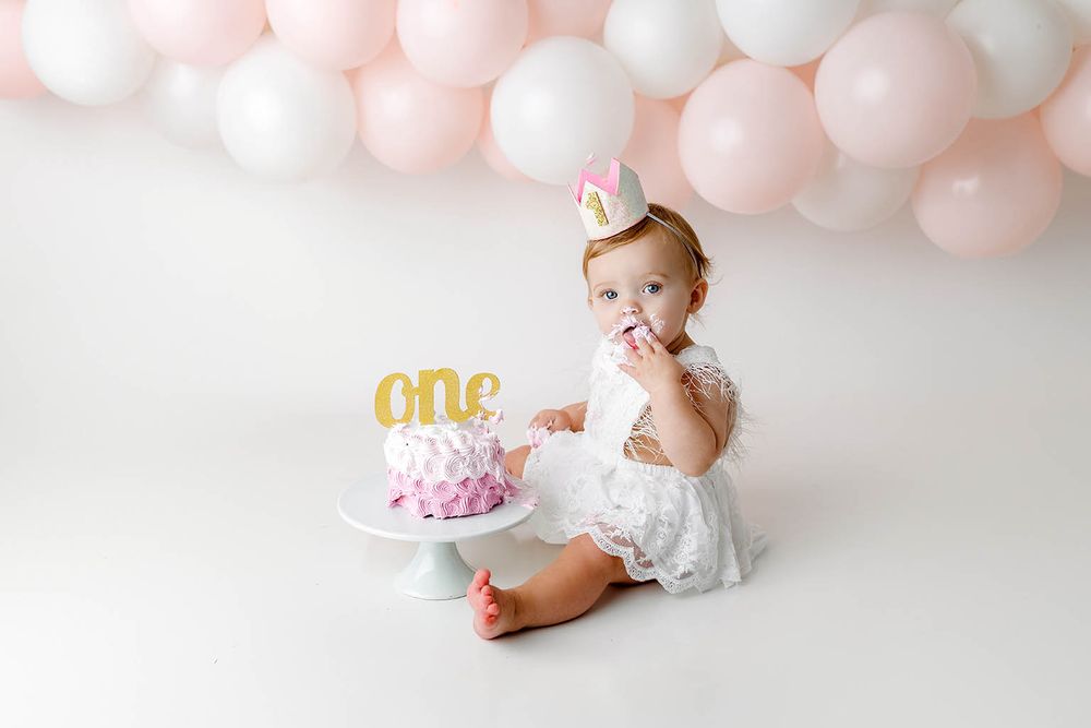 Adorable baby girl in white lace dress eating first birthday cake with gold one topper and pink balloon backdrop.