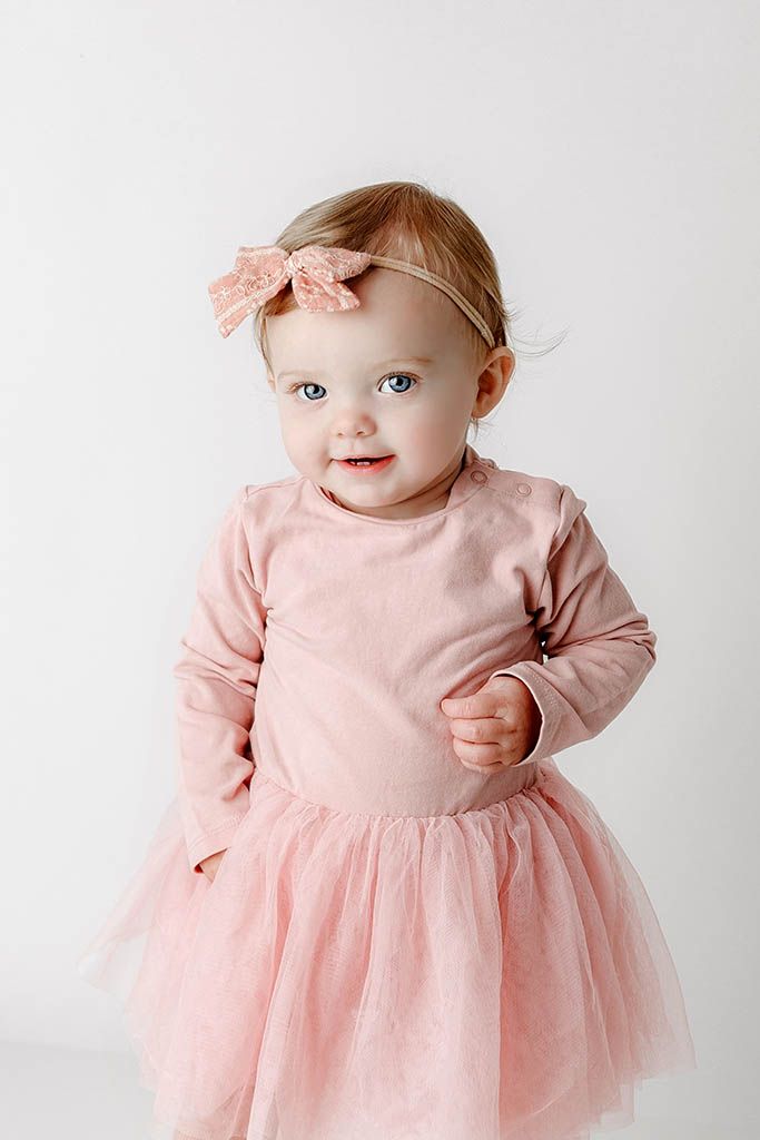 Adorable baby girl in pink tutu dress and bow headband smiling at camera against white background.