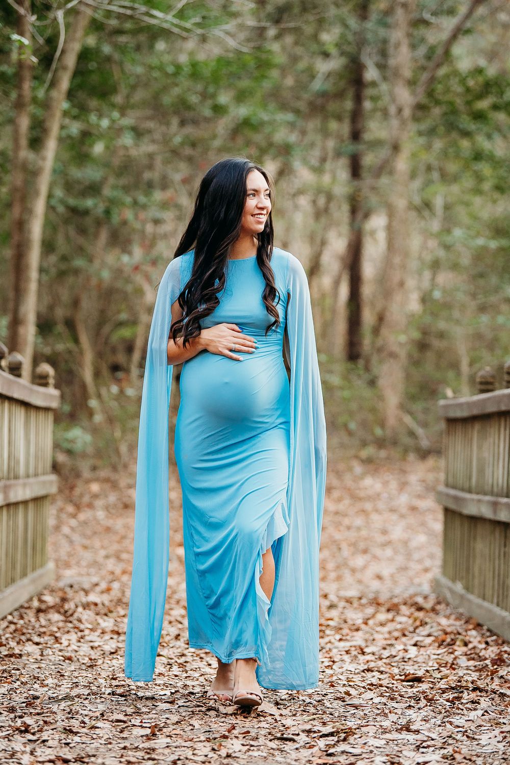 Beautiful pregnant woman in flowing light blue cape gown posing on wooden bridge surrounded by forest.