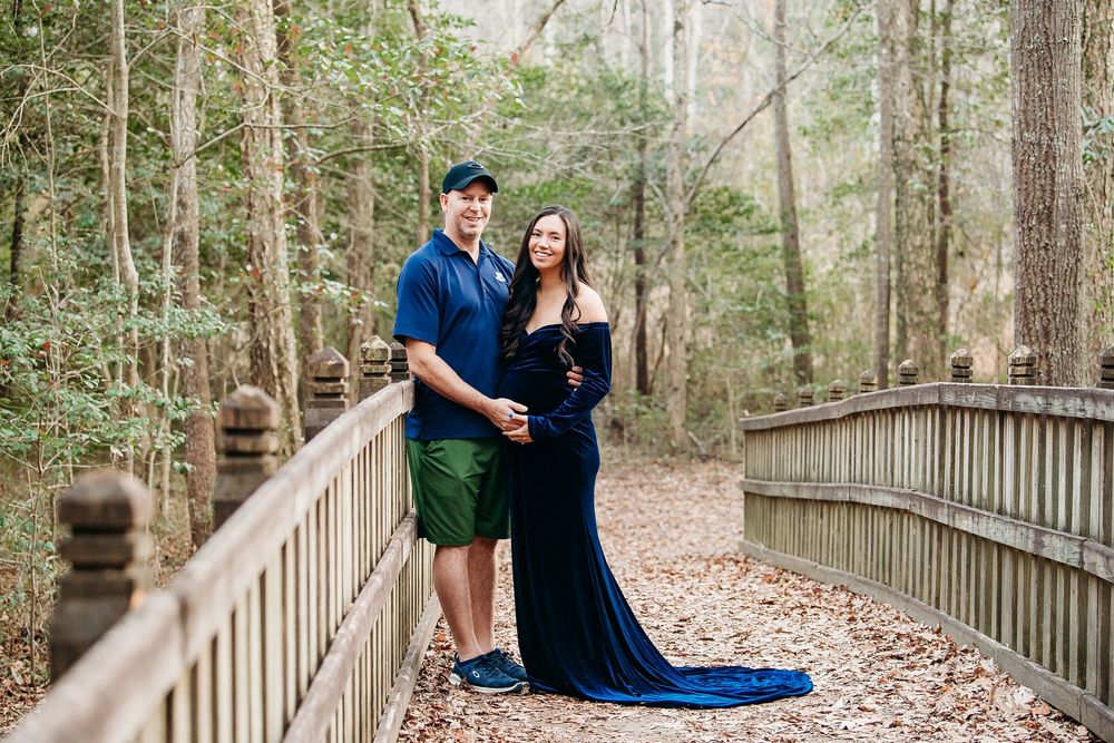 Couple poses on wooden bridge in forest, man in blue polo and woman in navy maternity gown.