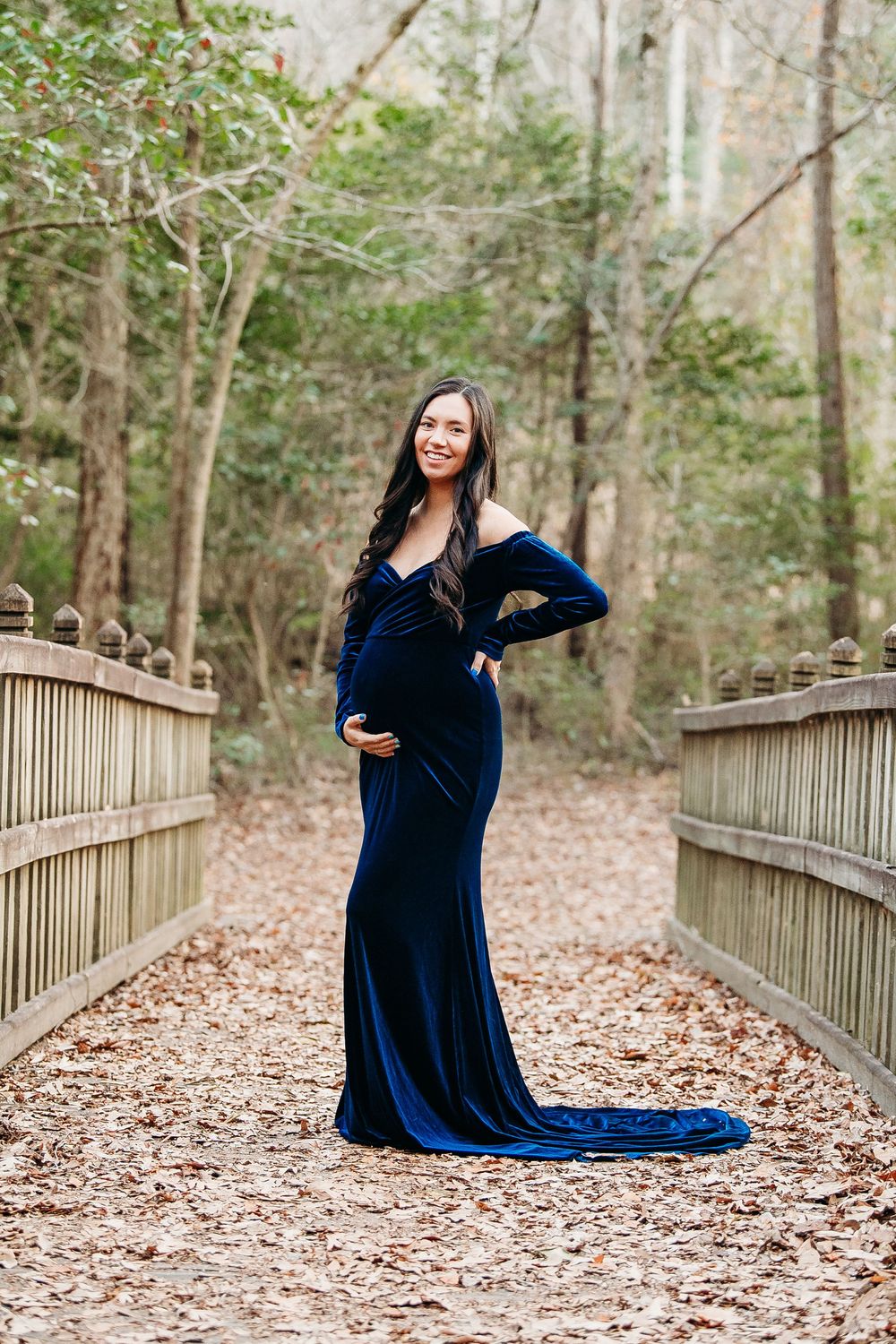 Pregnant woman in elegant navy blue velvet maternity gown poses on wooden bridge surrounded by trees.