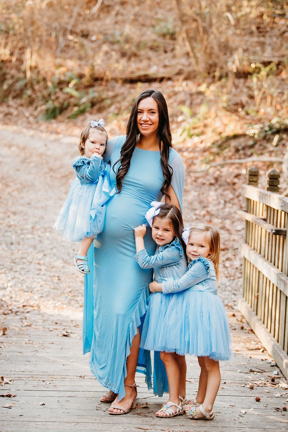 Pregnant mother in blue gown with three young daughters in matching blue dresses on wooden bridge.
