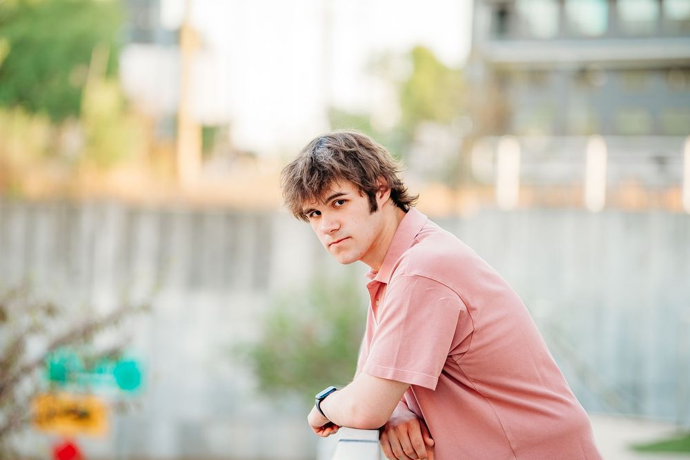 Young man in pink polo leaning on railing with blurred outdoor background.