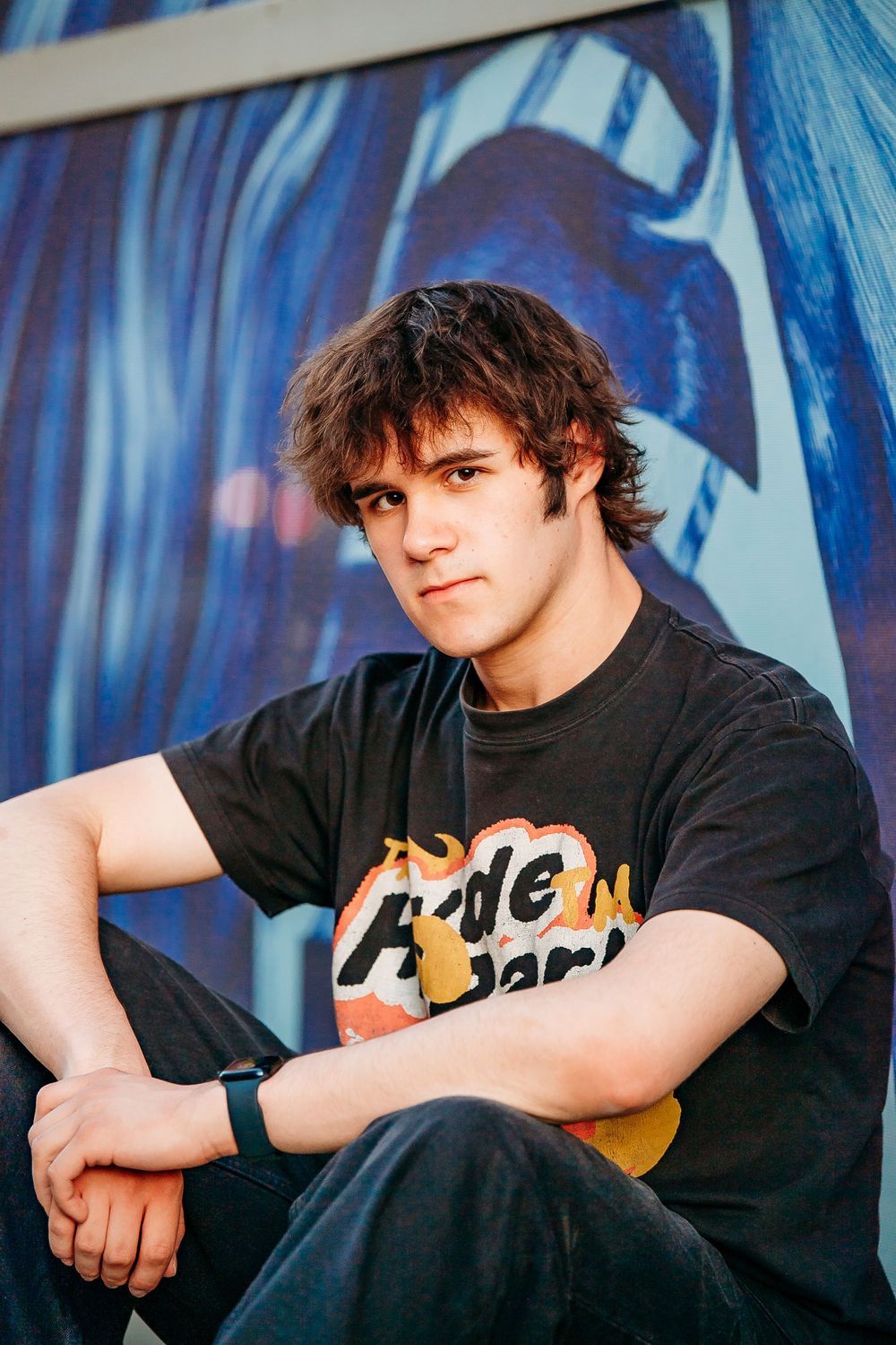 Teen boy in Hyde Park graphic tee sitting against colorful mural wall.
