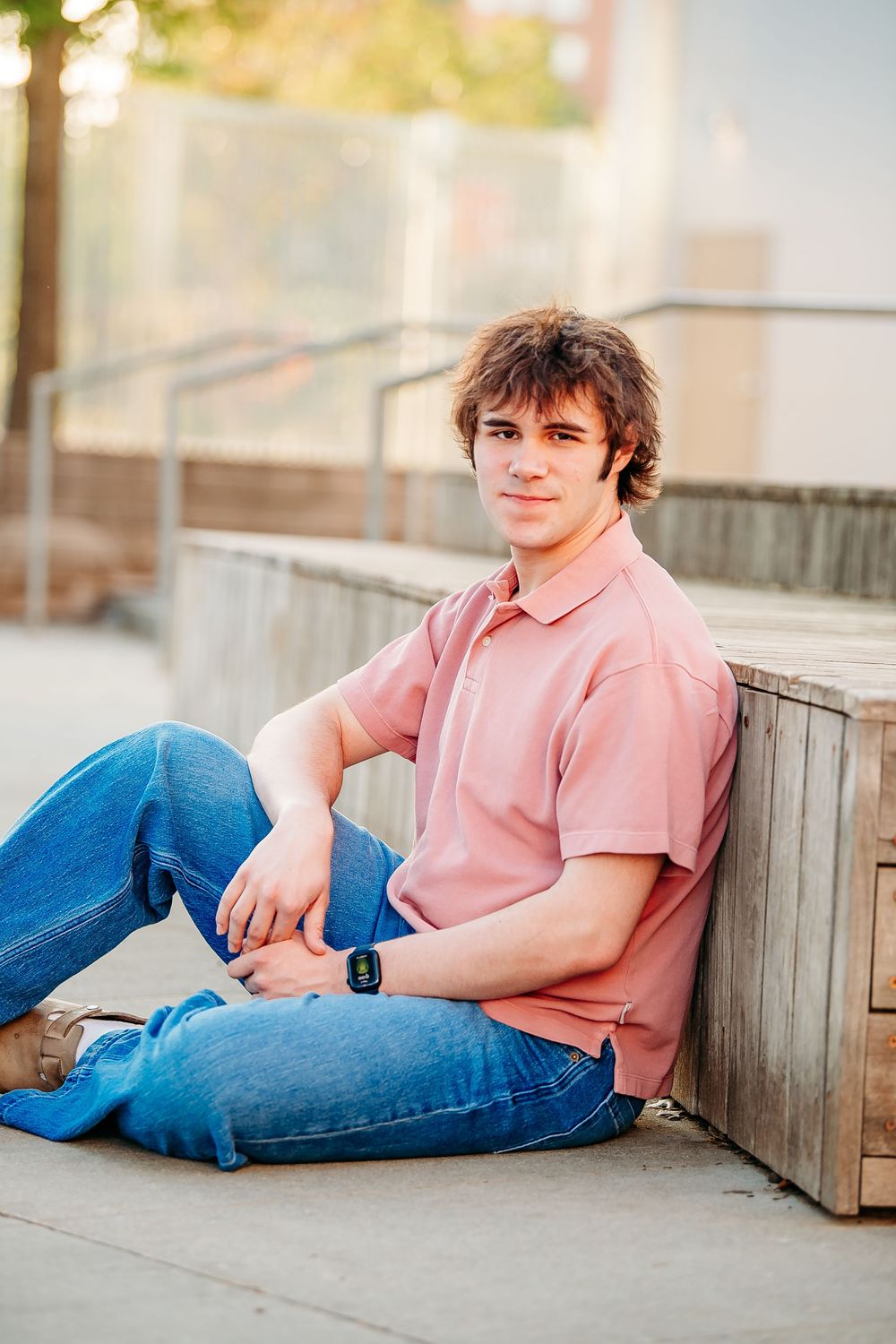Teen boy in pink polo and jeans sitting against wooden bench outdoors.