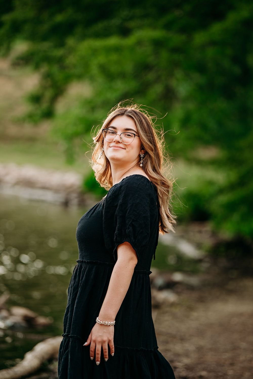 Young woman with glasses in black dress smiles with hair blowing near a scenic riverside.