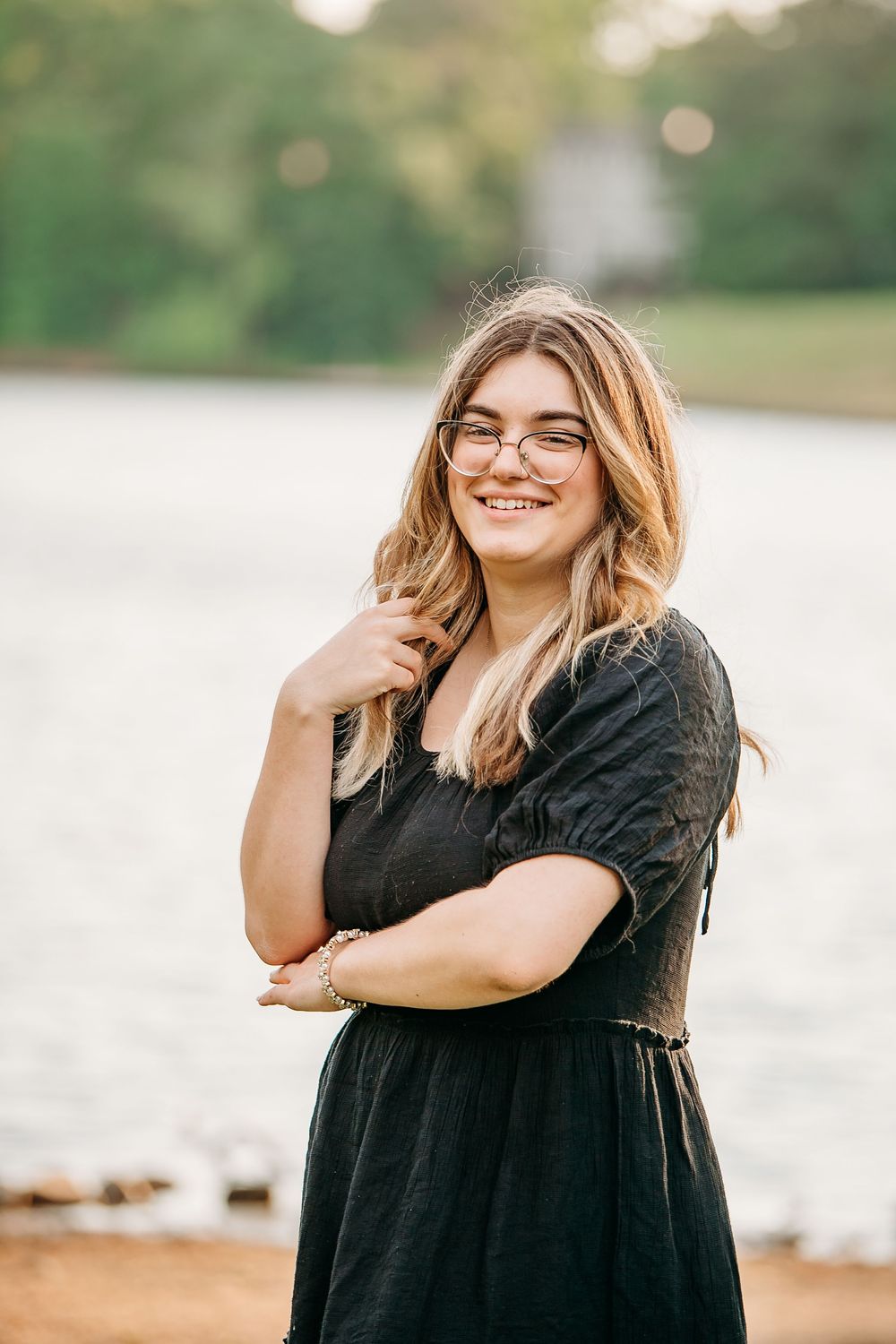 Smiling young woman with glasses in black dress poses by lakeside in warm natural light.
