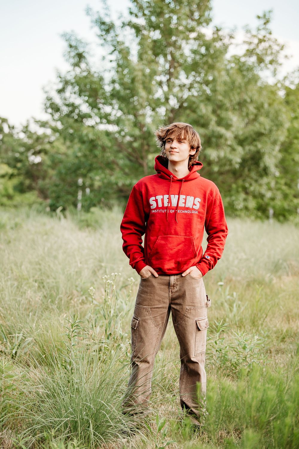 Teen boy in red Stevens Institute of Technology hoodie standing in tall grass field outdoors.