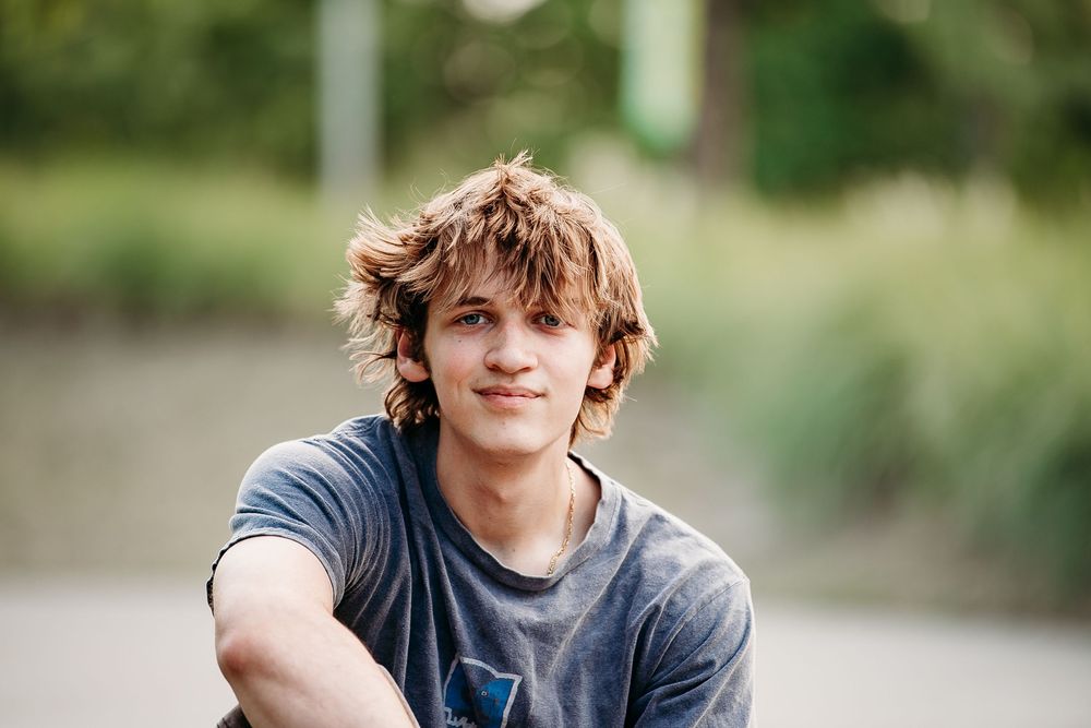 Smiling teenage boy with wavy brown hair wearing a gray t-shirt posing outdoors in natural light.