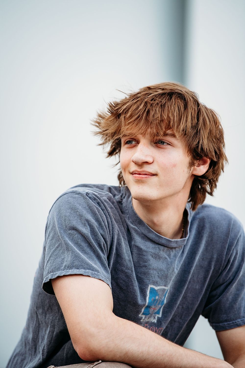 Young teen boy with tousled brown hair wearing a gray graphic tee sitting casually outdoors.