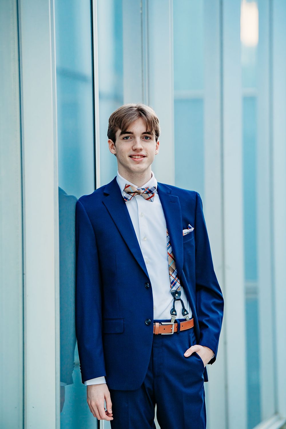 Dapper young man in navy blue suit and bow tie poses against modern glass wall backdrop.