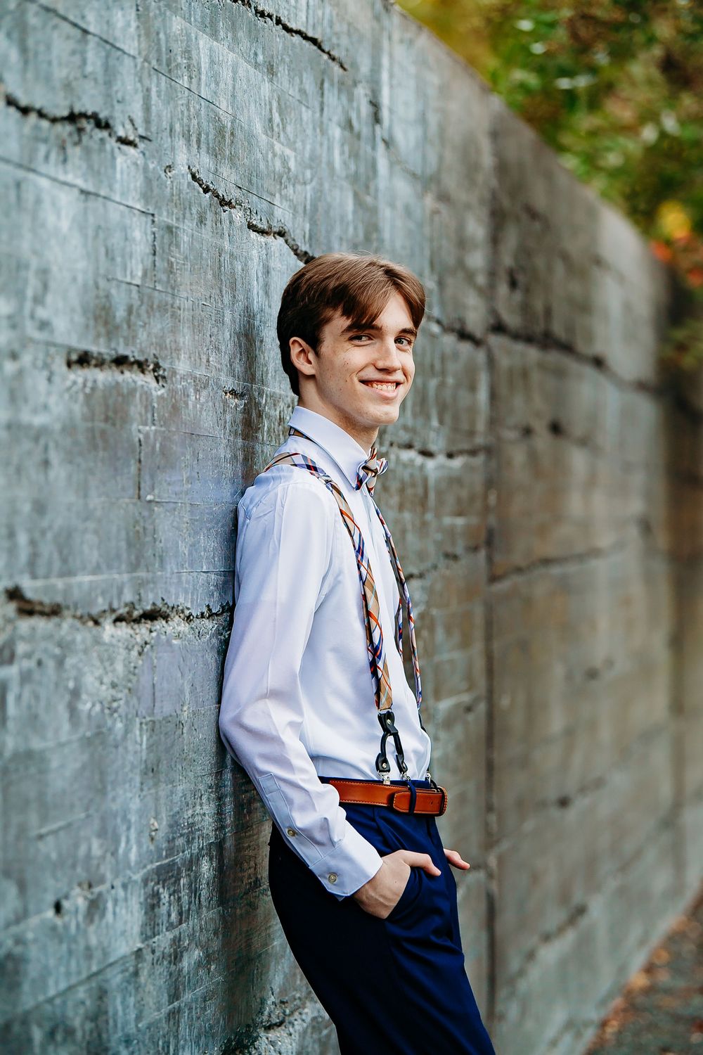 Young man in white shirt with suspenders smiles leaning against stone wall outdoors.