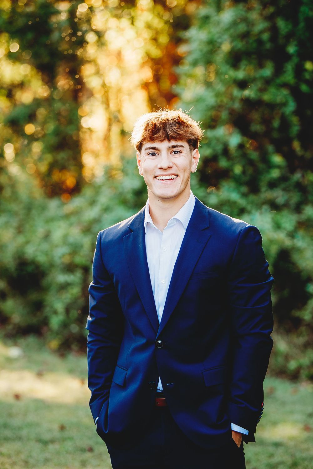 Smiling young man in navy blue suit stands outdoors in green park setting.