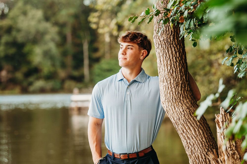 Young man in light blue polo leans against tree by serene lake in lush green outdoor setting.