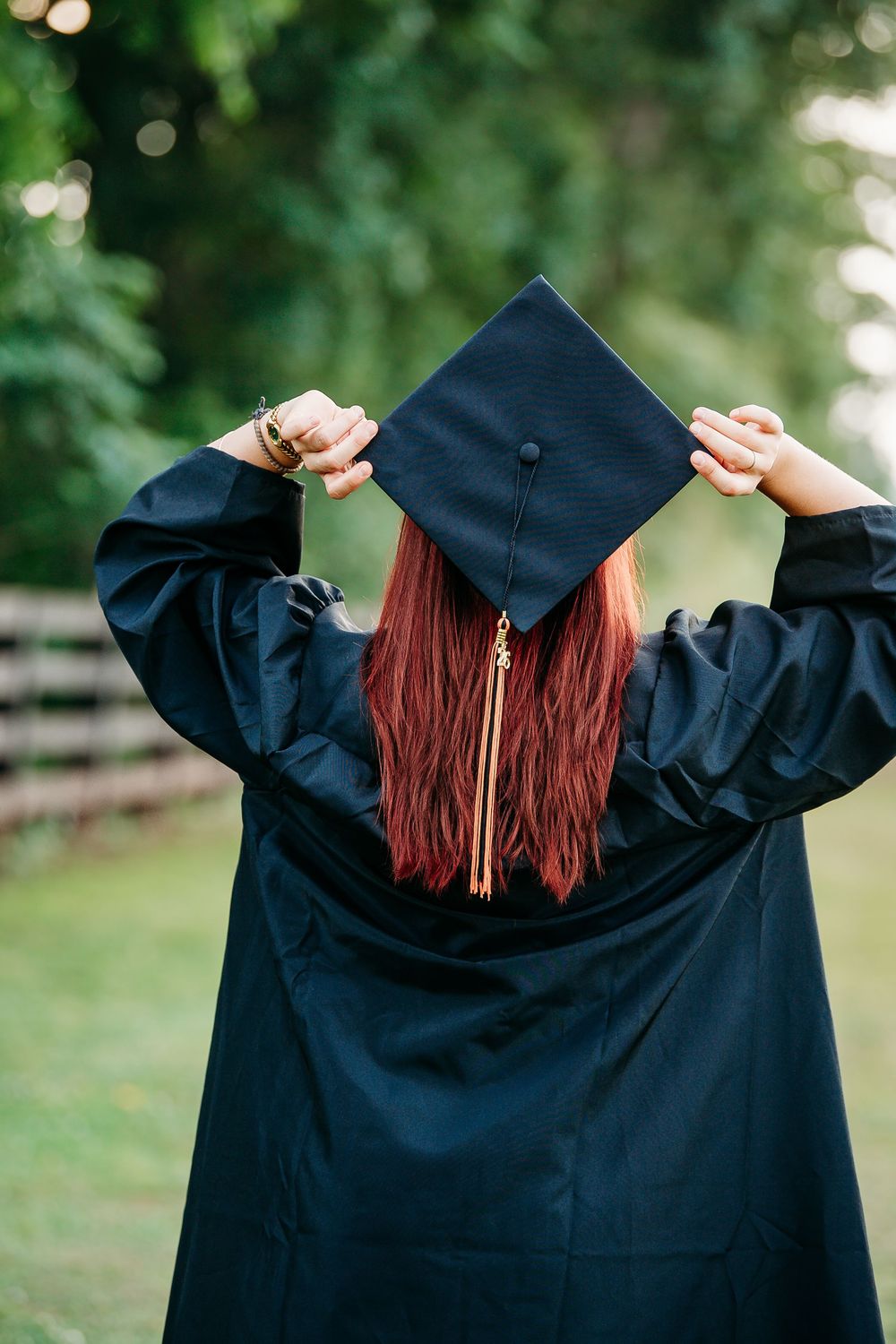 Graduate in black cap and gown holding mortarboard with red hair, viewed from behind outdoors.