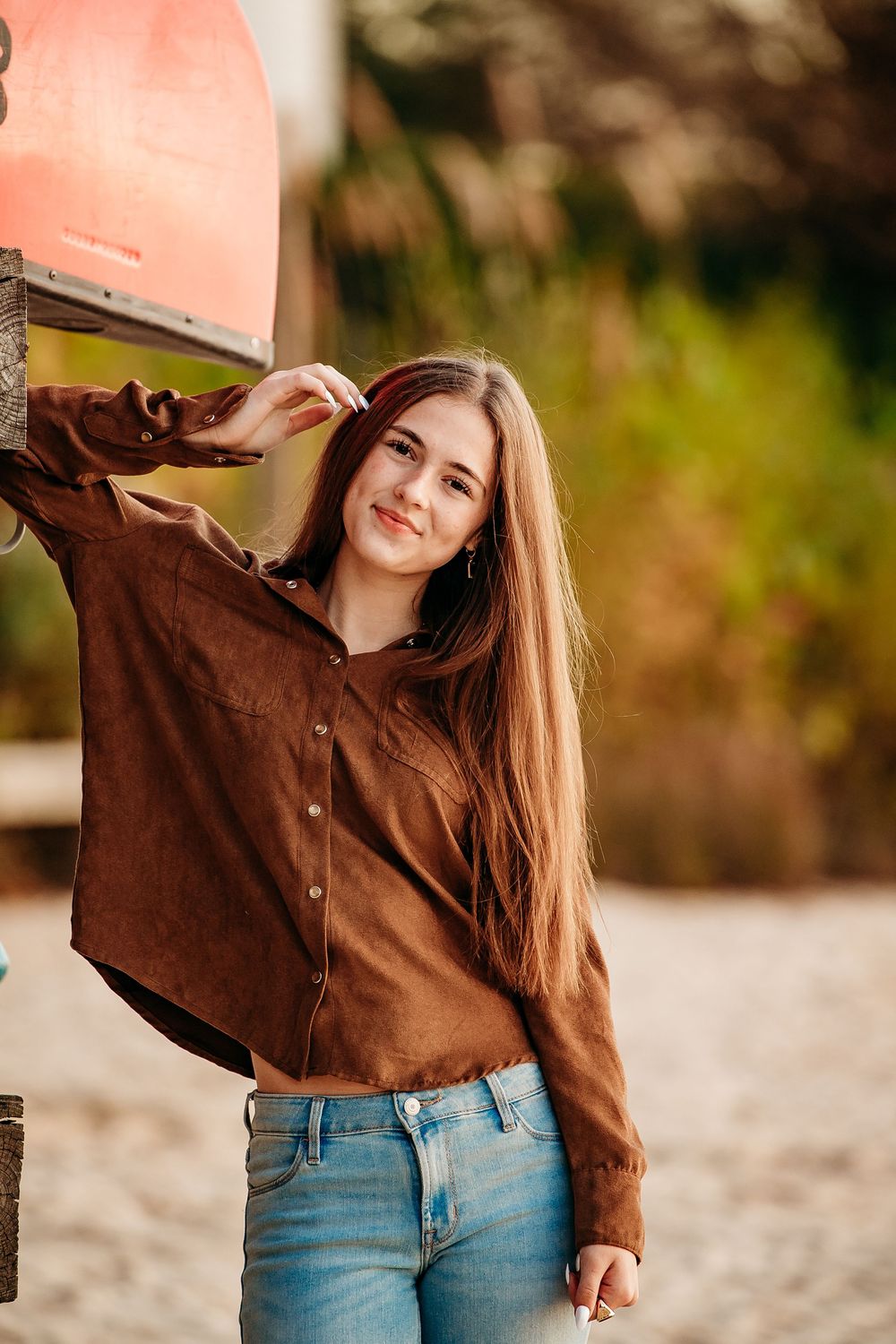Teen girl with long brown hair in brown suede shirt and jeans posing near a beach sign outdoors.