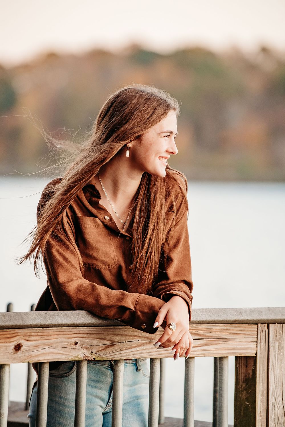 Smiling teenage girl with long brown hair leaning on wooden dock railing by a lake at sunset.