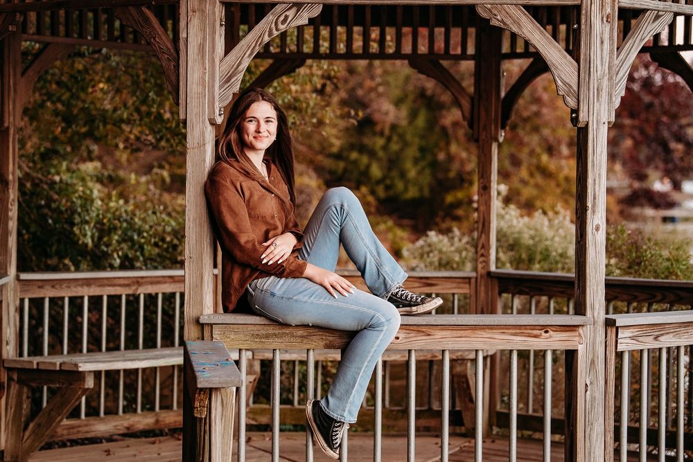 Smiling teenage girl in brown jacket and jeans sitting on wooden gazebo railing in autumn park.