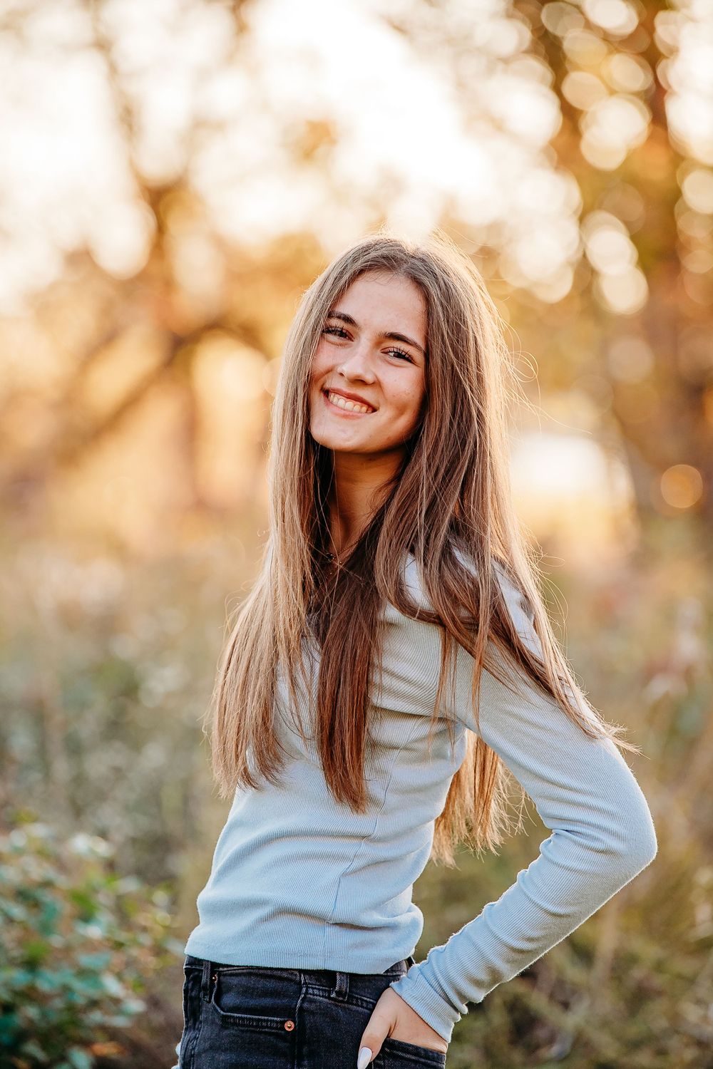 Happy teenage girl with long brown hair smiling in golden autumn sunlight wearing a light blue top.
