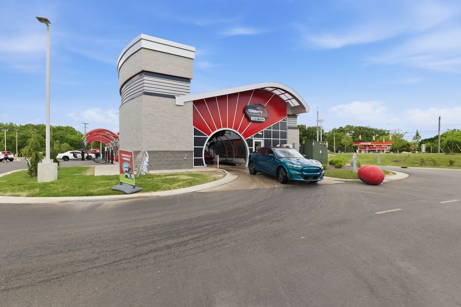 A teal car enters a modern red and gray car wash facility with a distinctive curved roof on a sunny day.