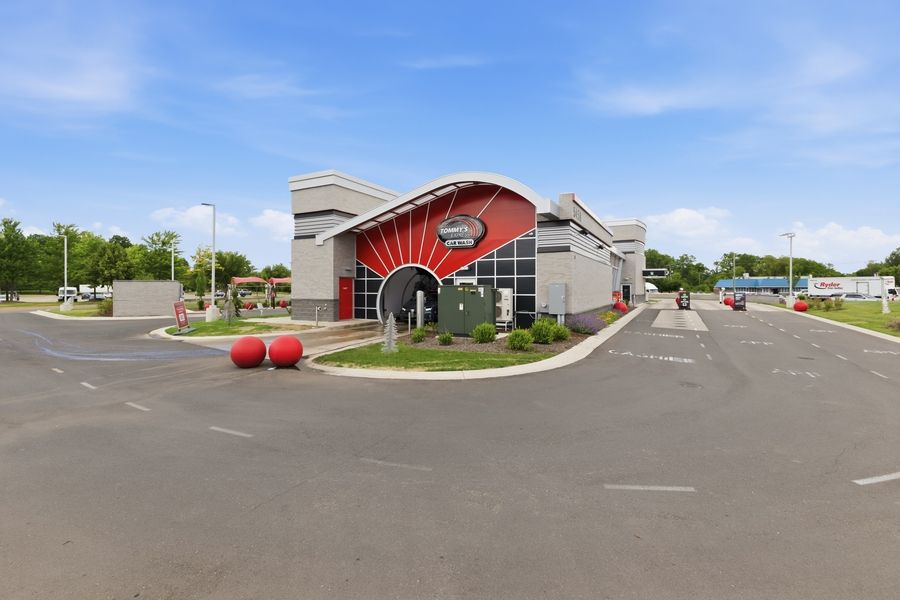 Modern red and gray commercial building with decorative sunburst facade, large parking lot, and lush green trees under blue sky.
