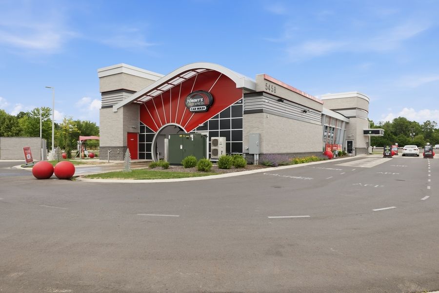 Modern commercial building with red arch entrance, large parking lot, and decorative red spheres on a sunny day.