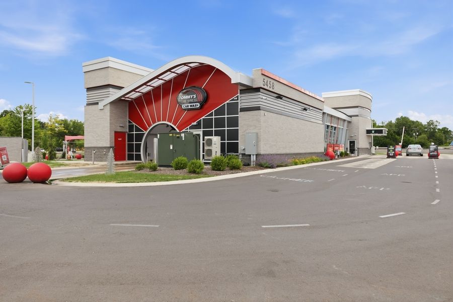 Exterior view of a modern commercial building with red accents and large parking lot under a clear blue sky.