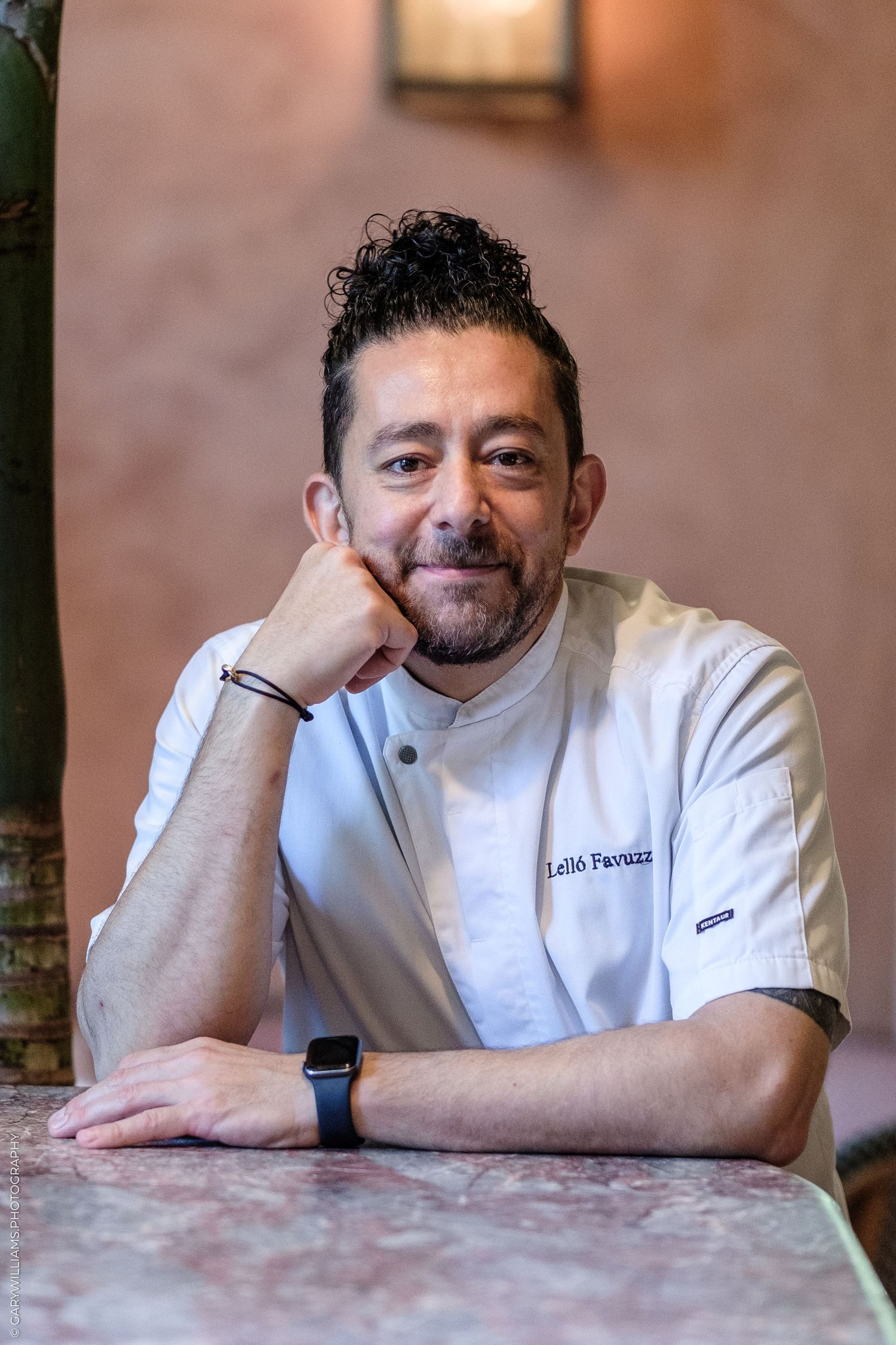 Chef in white uniform smiling warmly, leaning on marble table in elegant restaurant setting.