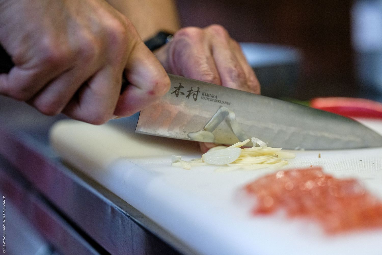 Chef finely mincing garlic on white cutting board with Japanese chef knife, diced tomatoes visible nearby.