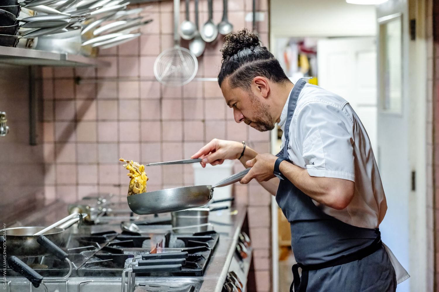Chef in apron sautéing pasta in a pan over a gas stove in a professional kitchen.