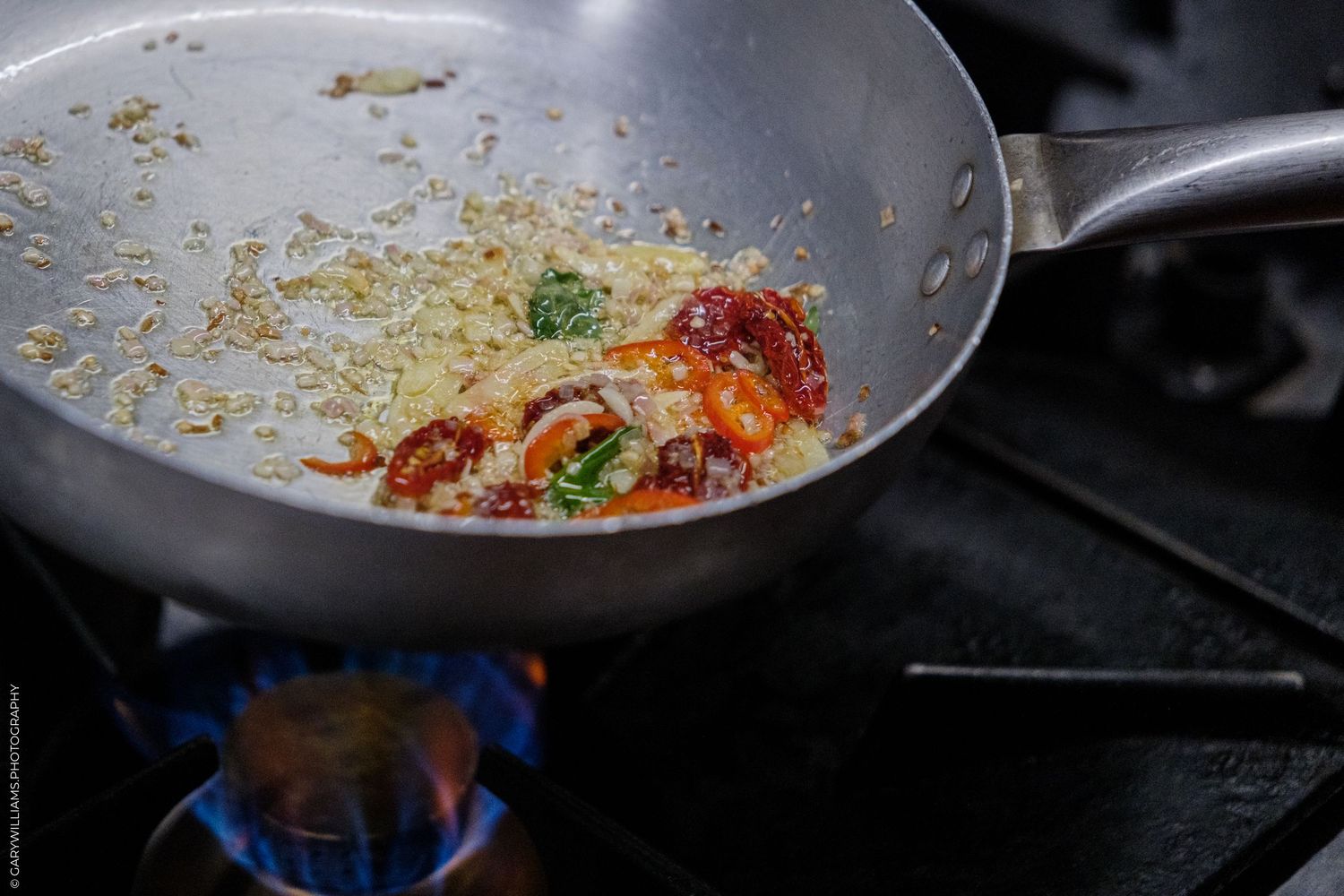 Sautéed vegetables with spices cooking in a stainless steel pan over a blue gas flame on a stovetop.