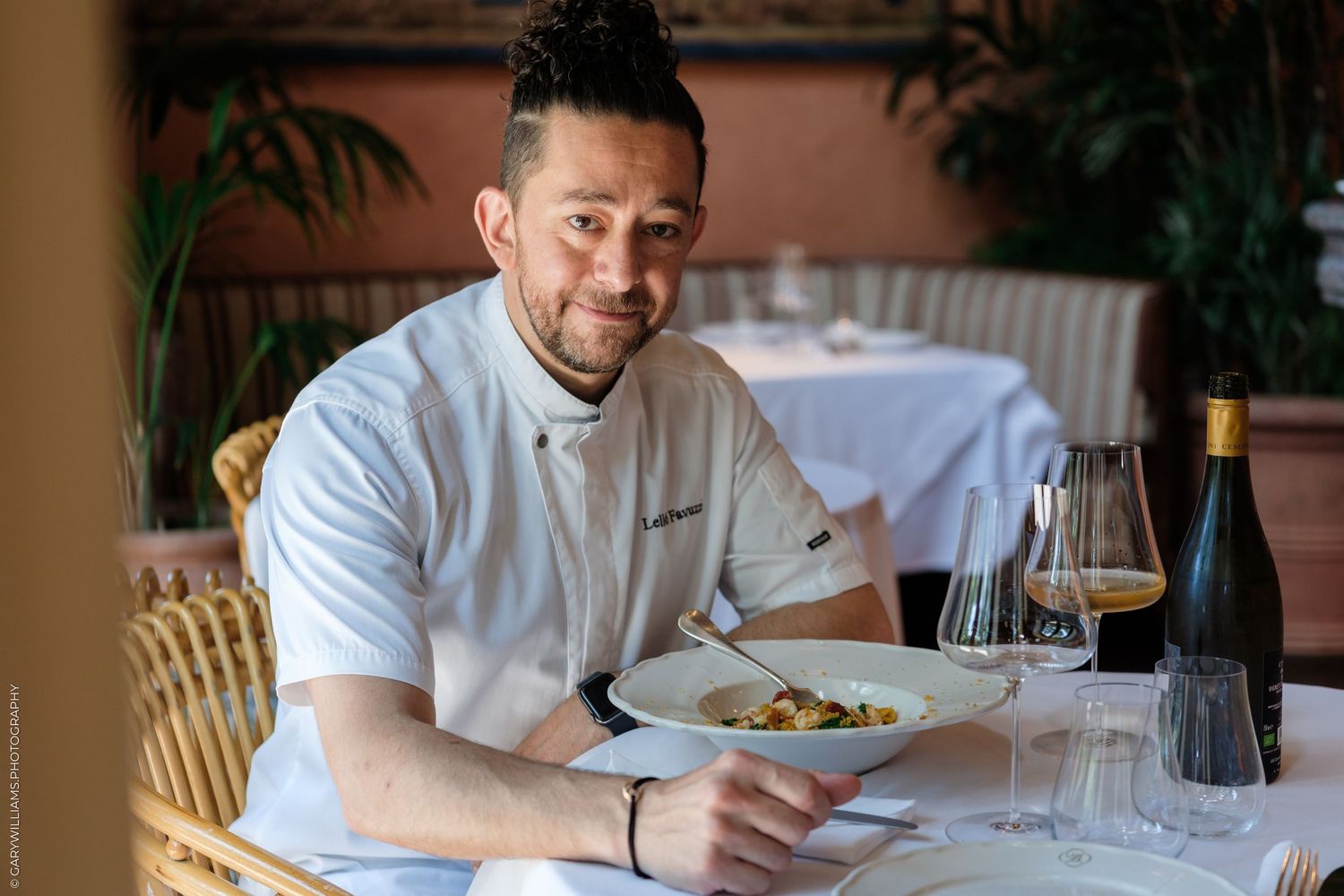 Smiling chef in white uniform presenting a gourmet pasta dish at an elegant restaurant table with wine glasses.