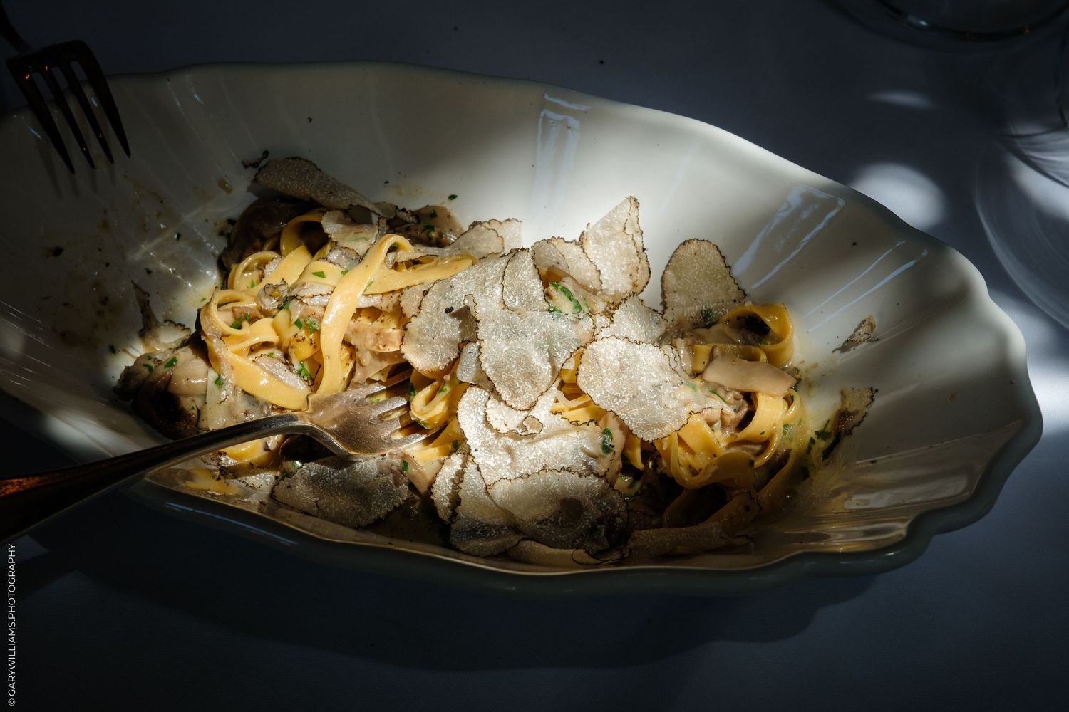 Elegant white bowl with fettuccine pasta topped with shaved truffles and mushrooms, served in dramatic lighting.