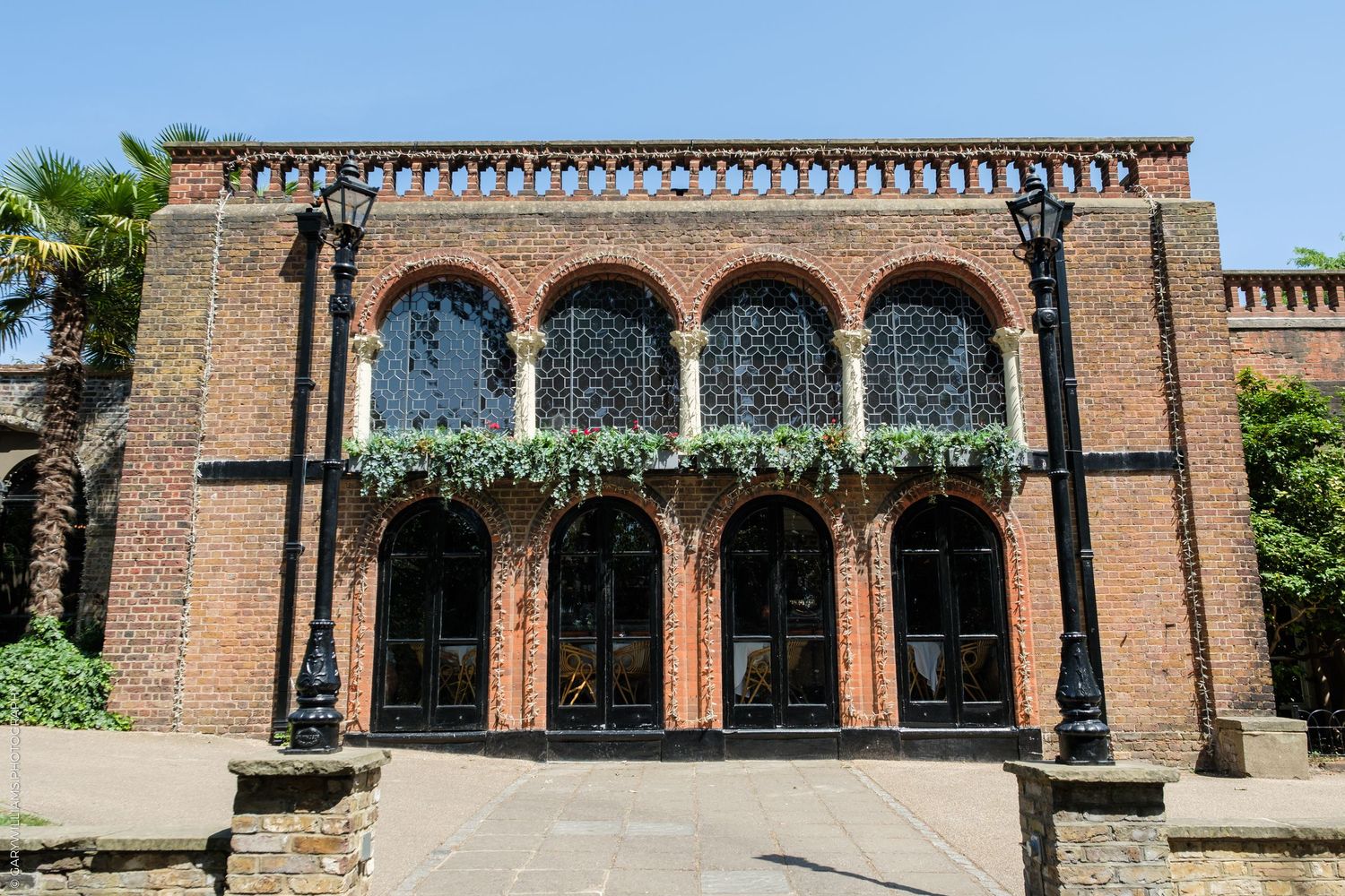Historic red brick building with arched windows, ornate black ironwork, and climbing ivy under a clear blue sky.