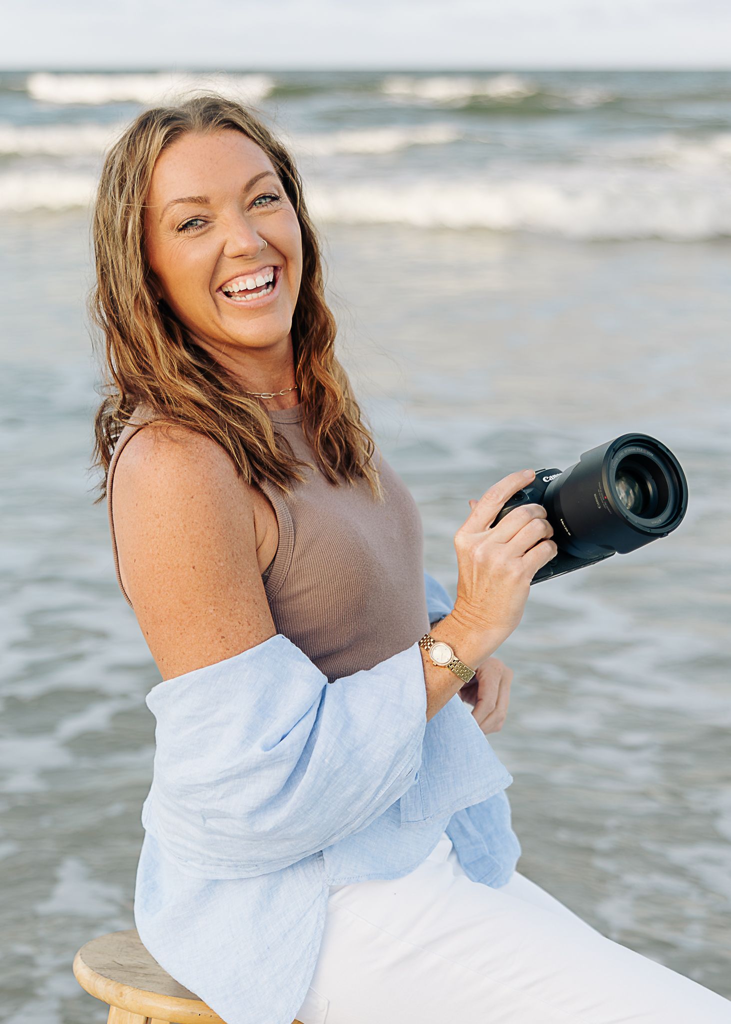 Woman in white jeans and blue shirt standing barefoot in ocean waves on a beach at sunset.